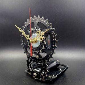 Könnte beinhalten: Eine einzigartige Uhr aus recycelten Fahrradteilen. Die Uhr hat ein schwarzes Zahnrad als Zifferblatt, goldfarbene Zeiger und einen roten Sekundenzeiger. Sie ist auf einem schwarzen Sockel montiert und schafft ein industrielles und künstlerisches Design.