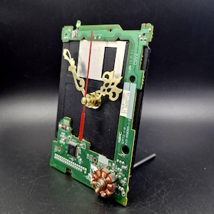 Puede incluir: Un reloj hecho de una placa de circuito con un segundero rojo y agujas de hora y minuto doradas. El reloj está montado en un soporte negro.
