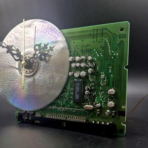 Op de afbeelding: Een wandklok gemaakt van een groene rechthoekige printplaat en een ronde zilveren compact disc. De klok is voorzien van sierlijke zwarte metalen wijzers, een dunne gouden secondewijzer en diverse elektronische componenten.
