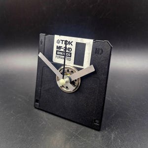 May include: A black TDK MF-2HD IBM/DOS formatted floppy disk repurposed as a clock. The square disk features a silver metal shutter and a central clock mechanism with two silver-colored hands and a thin black second hand attached to the front.