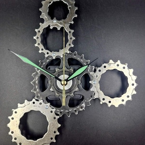 Könnte beinhalten: Eine Wanduhr aus silberfarbenen Fahrradzahnrädern, die in einer Gruppe angeordnet sind. Die Uhr verfügt über ein zentrales Uhrwerk mit zwei hellgrünen, spitzen Zeigern und einem dünnen goldfarbenen Sekundenzeiger vor dunklem Hintergrund.