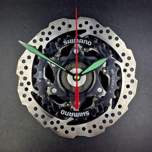 May include: A wall clock constructed from a circular silver metal bicycle brake rotor and a black Shimano MF-TZ500-7 gear cassette. The clock features two light green hour and minute hands and a thin red second hand centered on the mechanism.
