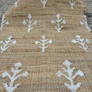 Può includere: Un tappeto in iuta color naturale con un motivo floreale bianco ripetuto. Il tappeto ha una trama tessuta e un bordo arrotolato, che mostra una miscela di toni neutri e un design semplice ma elegante. Adatto a vari stili di interni.