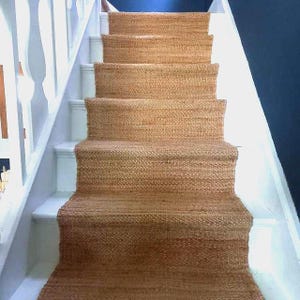 Può includere: Una passatoia per scale tessuta di colore marrone naturale copre i gradini di una scala. La passatoia è realizzata in un materiale strutturato ed è installata su scale verniciate di bianco con una balaustra bianca sul lato sinistro. Il muro è dipinto di blu scuro.