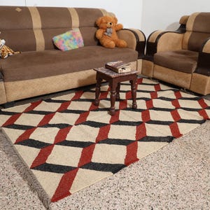 May include: A living room scene with a brown sofa and armchair with tan detailing. A geometric rug with red, black, and cream cube patterns is on the floor. A small wooden table and a potted plant complete the scene.