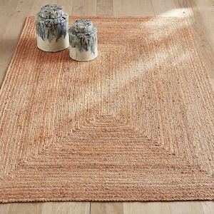 Puede incluir: Una alfombra rectangular tejida en color tostado natural con una textura trenzada visible. Dos recipientes cilíndricos de cerámica con tapa están colocados sobre la alfombra, con una base blanca y un patrón de esmalte azul grisáceo arriba.