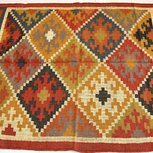 Puede incluir: Una alfombra colorida con estampado geométrico en un esquema de colores rojo, naranja, amarillo, marrón y negro. La alfombra tiene un borde de flecos y está hecha de yute tejido.