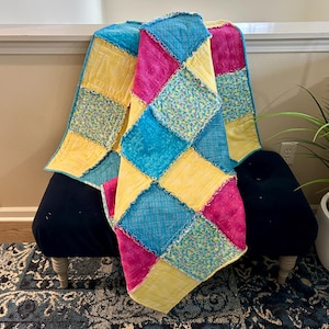 May include: A colorful patchwork quilt with squares of yellow, pink, blue, and green fabric. The quilt is made with a variety of patterns and textures, and has a frayed edge.