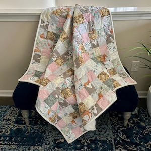 Puede incluir: Una colcha de retazos con un borde blanco y una variedad de patrones que incluyen ciervos, diseños florales y formas geométricas en tonos de rosa, marrón, beige y verde.