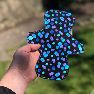 May include: A black reusable cloth menstrual pad with a blue and purple polka dot pattern.