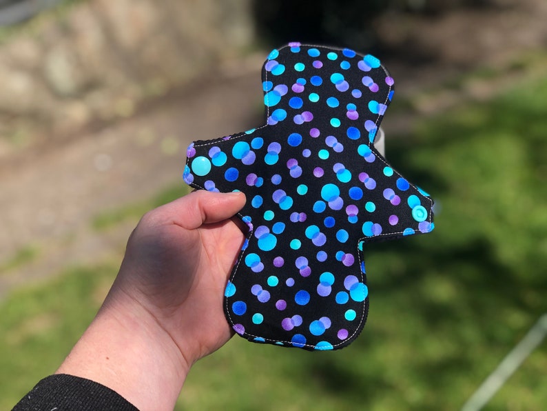 May include: A black reusable cloth menstrual pad with a blue and purple polka dot pattern.