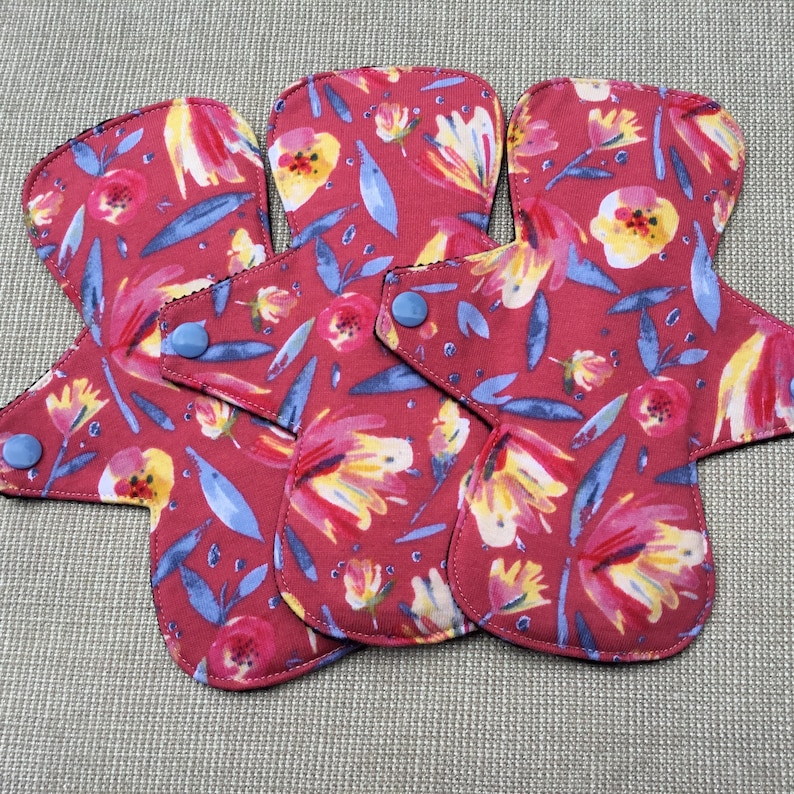 May include: Three reusable cloth menstrual pads with a floral print in shades of red, yellow, blue, and white. The pads have snaps on the sides for attaching to underwear.