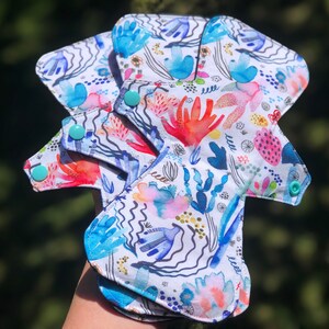 May include: Three reusable cloth menstrual pads with a colorful abstract floral print. The pads have turquoise snaps and a white background.