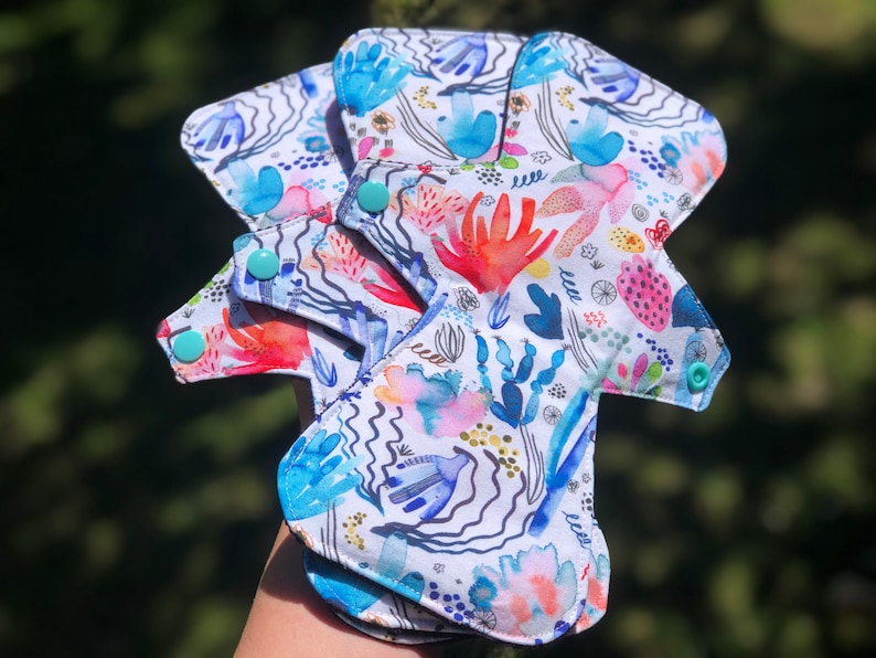 May include: Three reusable cloth menstrual pads with a colorful abstract floral print. The pads have turquoise snaps and a white background.