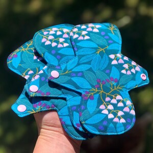 May include: Three reusable cloth menstrual pads with a floral print in shades of teal, green, and purple. The pads have white snaps and are folded together.