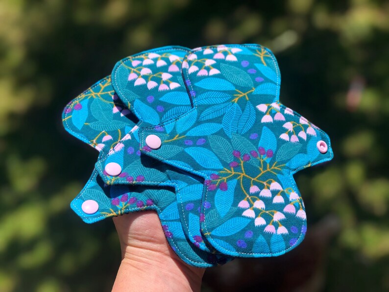 May include: Three reusable cloth menstrual pads with a floral print in shades of teal, green, and purple. The pads have white snaps and are folded together.