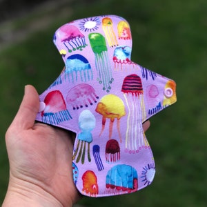 May include: A purple reusable cloth pad with a colorful jellyfish print. The pad has two white snaps on the sides.