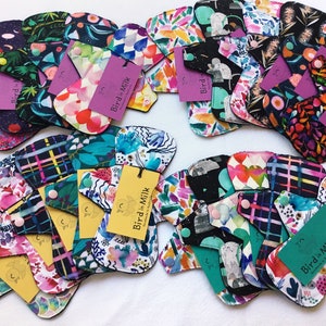 May include: A variety of colorful reusable cloth menstrual pads with a black trim. The pads feature different floral and abstract patterns. The pads are labeled with the text "Bird in Milk".