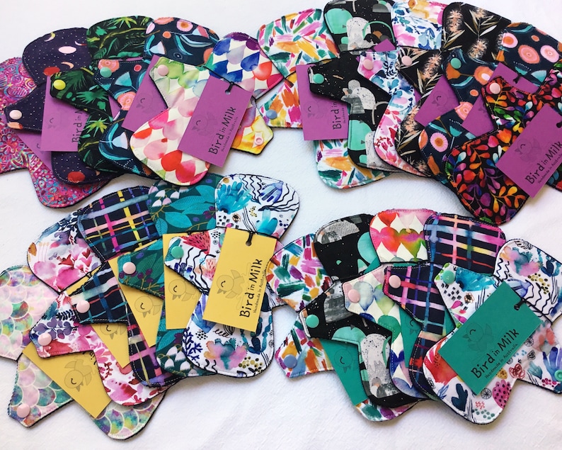 May include: A variety of colorful reusable cloth menstrual pads with a black trim. The pads feature different floral and abstract patterns. The pads are labeled with the text "Bird in Milk".