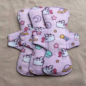 May include: Three reusable cloth pads with a pink background and a repeating pattern of a cartoon cat with wings, stars, moons, and planets. The pads have pink snaps on the sides.