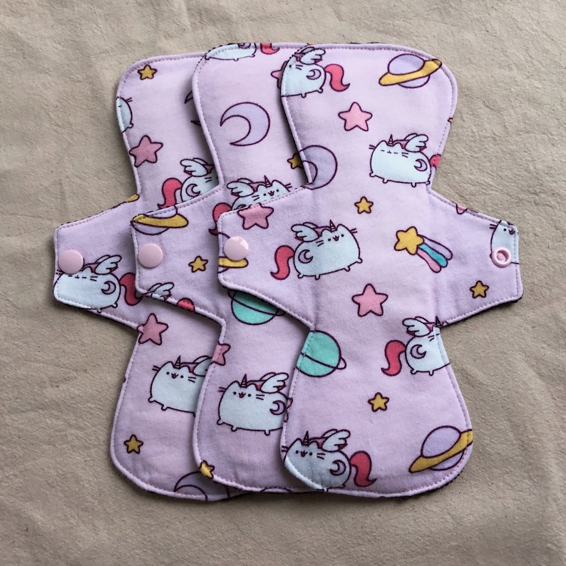 May include: Three reusable cloth pads with a pink background and a repeating pattern of a cartoon cat with wings, stars, moons, and planets. The pads have pink snaps on the sides.