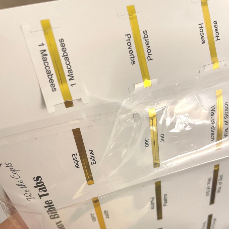 May include: Close-up of gold Bible tabs with the names of books of the Bible printed on them, including Esther, Job, Proverbs, and 1 Maccabees.