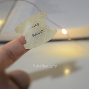 May include: A close-up of a finger holding a yellow sticker with the word "Baruch" written on it. The sticker has a grid pattern and is slightly torn. The background is blurry and features a string of lights.