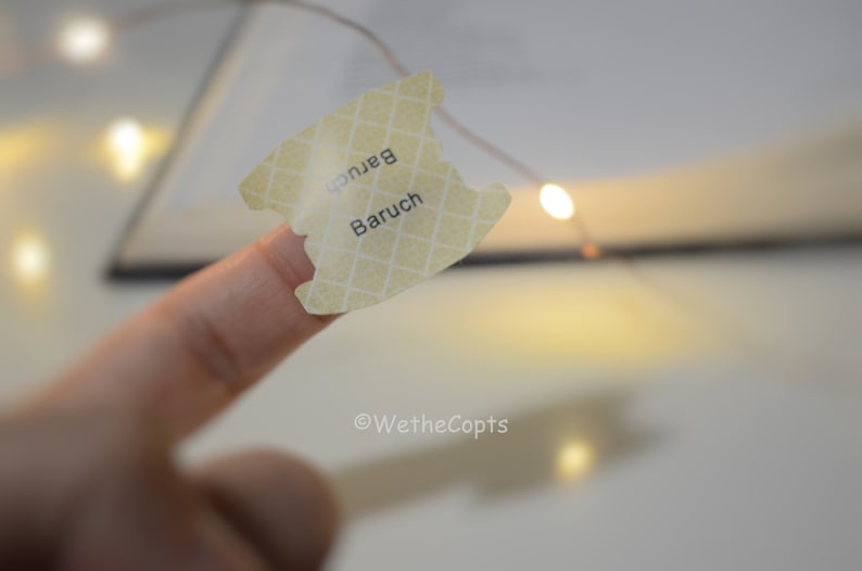 May include: A close-up of a finger holding a yellow sticker with the word "Baruch" written on it. The sticker has a grid pattern and is slightly torn. The background is blurry and features a string of lights.