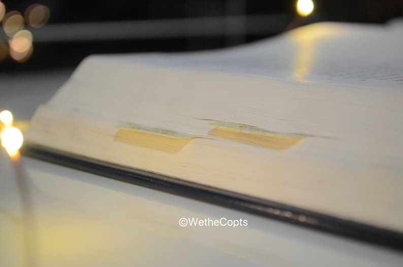 May include: Close-up of a thick, white book with a blue spine. The book has two yellow bookmarks sticking out of the pages. The book is on a white surface.
