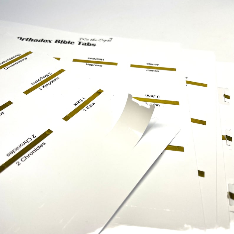 May include: A set of gold colored Bible tabs with the names of books of the Bible printed on them, including Deuteronomy, 2 Kingdoms, 1 Ezra, 2 Chronicles, Hebrews, James, and 3 John. The tabs are labeled "We the Copts Orthodox Bible Tabs".
