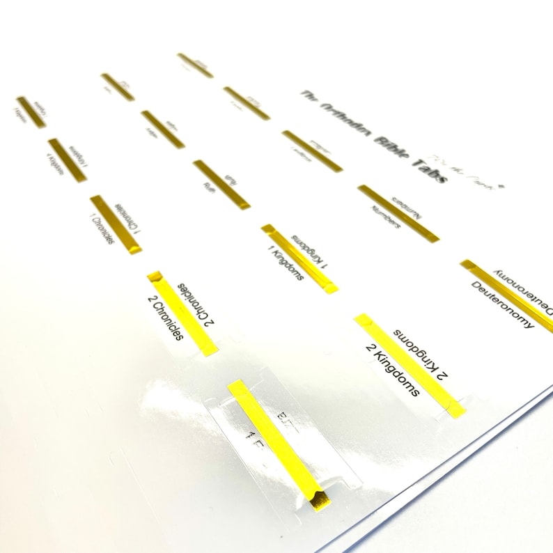 May include: A set of yellow and gold Bible tabs with the names of books of the Bible printed on them. The tabs are labeled with the names of books from the Old Testament, including Deuteronomy, Numbers, 1 Kings, 2 Kings, 1 Chronicles, 2 Chronicles, and Ezra.