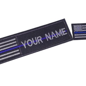 以下が含まれることがあります： アメリカの国旗に薄い青色の線が引かれ、「YOUR NAME」という文字が書かれた黒と灰色の刺繍パッチです。