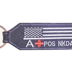 May include: A black and white American flag keychain with the text "A+ POS NKDA" in white. The keychain has a silver ring.