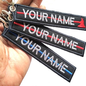 May include: Three black keychains with a thin red, blue, or red and blue line and the text "YOUR NAME" in white. The keychains are held in a hand.