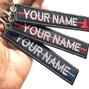 May include: Three black keychains with a thin red, blue, or red and blue line and the text "YOUR NAME" in white. The keychains are held in a hand.
