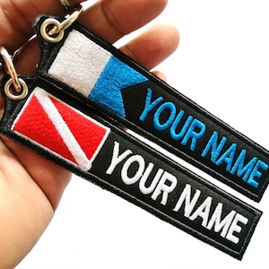 Puede incluir: Dos llaveros de tela negra con bordados blancos y azules. Un llavero tiene una bandera de buceo y el texto "YOUR NAME" en blanco. El otro llavero tiene una bandera azul y blanca y el texto "YOUR NAME" en azul.