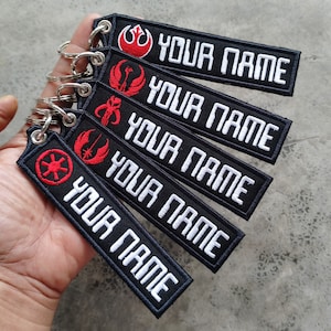 Puede incluir: Cinco llaveros negros con texto blanco que dice "YOUR NAME" y diferentes símbolos de Star Wars en rojo. Los símbolos son la Alianza Rebelde, el símbolo mandaloriano, la Orden Jedi, el Imperio Galáctico y la Alianza Rebelde.