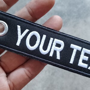 May include: Black fabric keychain with a silver metal ring and a white embroidered text that says "YOUR TEXT".