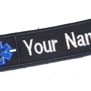 May include: A black embroidered patch with the text "Your Name" in white. The patch features a blue Star of Life emblem on the left side, commonly associated with emergency medical services.