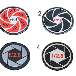 Op de afbeelding: Vier ronde zwart-witte camera-diafragma-patches met rode tekst. De patches zijn genummerd van 1 tot en met 4. Patch 1 heeft de tekst "f/1.4", "f/2.8", "f/4", "f/5.6", "f/8", "f/16". Patch 2 heeft de tekst "f/2.8". Patch 3 heeft de tekst "f/2.8". Patch 4 heeft de tekst "f/2.8".