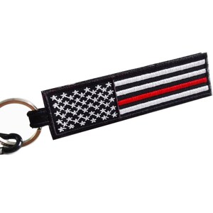 May include: Black keychain with a red line through the middle of an American flag. The keychain has a silver metal ring and a silver carabiner clip.