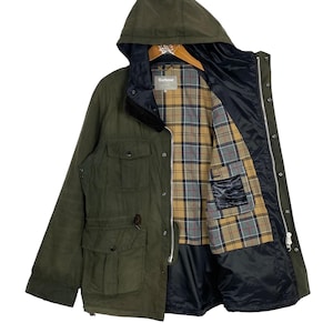 Pode incluir: Uma jaqueta Barbour verde com capuz e forro xadrez. A jaqueta tem fecho de correr e vários bolsos.