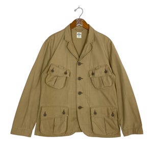 May include: A tan-colored jacket with multiple front pockets and button closures. The jacket has a collared neckline and long sleeves. It is hanging on a wooden hanger.