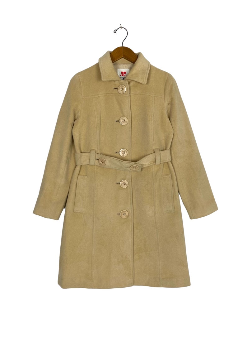 May include: A beige belted coat with a collared neckline and button closure. The coat has a classic design and is made of a soft, warm fabric.