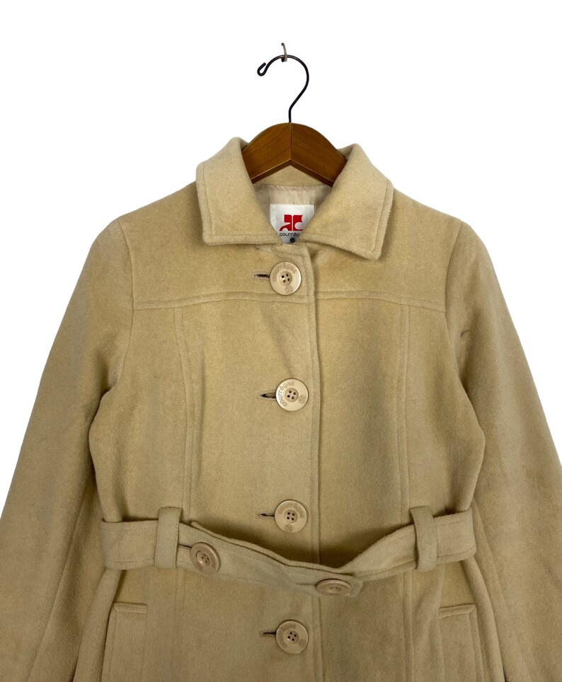 May include: A beige wool coat with a belt and five large buttons. The coat has a collar and long sleeves. The label on the coat reads "JC Collection".