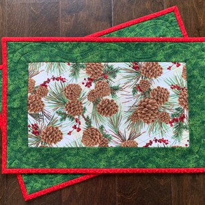 May include: Two quilted table runners with a pine cone and holly berry print. The runners have a green border with a red trim.