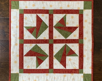 May include: A quilted wall hanging with a geometric design featuring red, green, and white fabric squares. The fabric has a subtle pattern of stars and swirls.