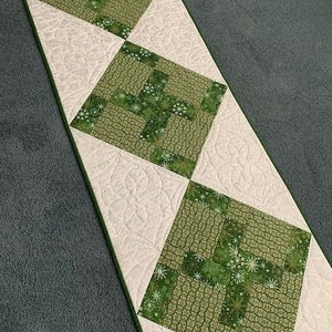 May include: A white quilted table runner with a green and white patterned design. The pattern features a repeating design of a cross with a floral motif.