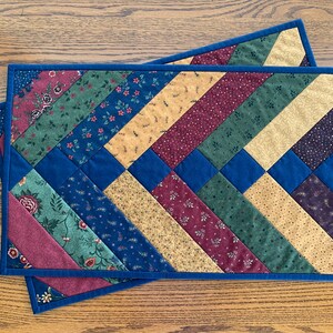May include: Two quilted placemats with a diagonal pattern of burgundy, green, gold, and blue fabric. The placemats have a dark blue border.