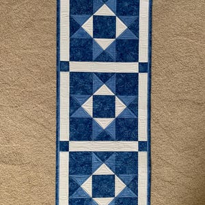 Puede incluir: Un camino de mesa acolchado rectangular con un diseño geométrico. El camino presenta un patrón de cuadrados y triángulos azules, con bordes y acentos blancos. La tela tiene un estampado floral. El camino mide aproximadamente 91 cm de largo y 30 cm de ancho.